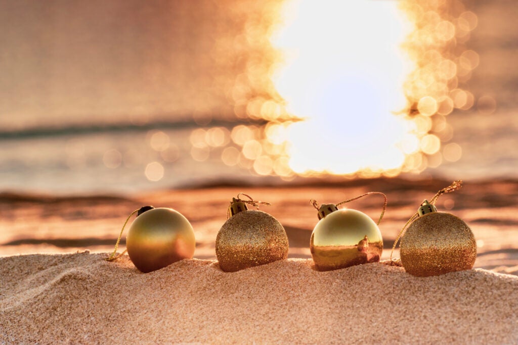 4 gold Christmas tree ornaments in the sand on a beach at sunset with ocean behind