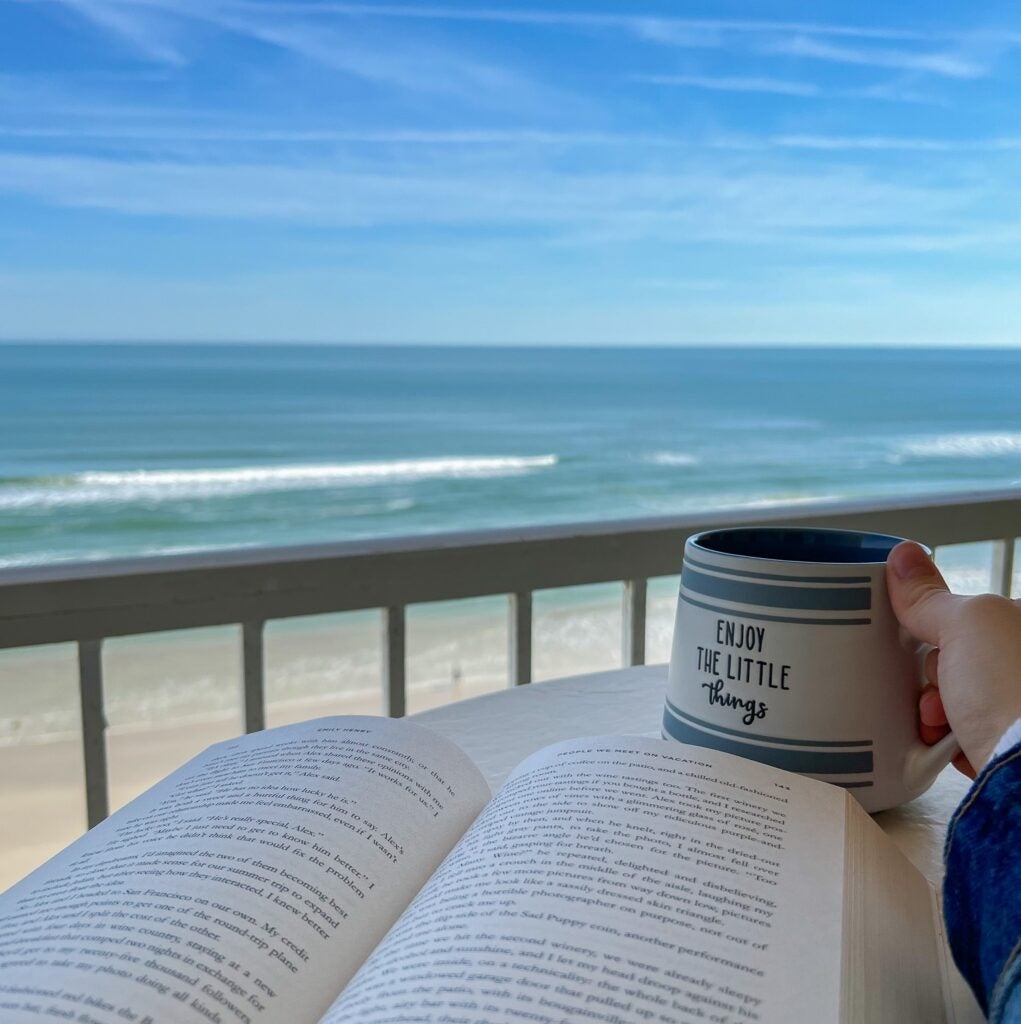 person sitting on oceanfront balcony with incredible view of teal ocean and blue sky, open book on table with hand holding mug that says enjoy the little things